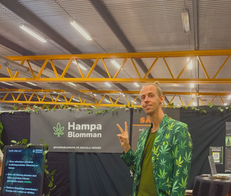 Man i grön hampablads-kavaj gör fredstecken framför Hampablommans banderoll på Harmoni Expo 2025